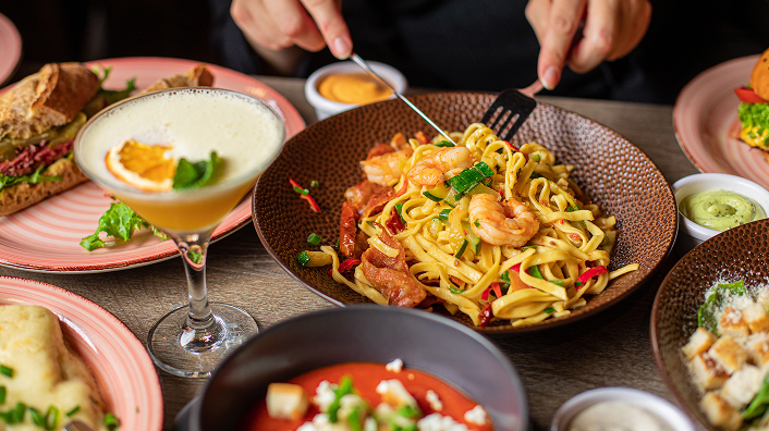 A person sitting at a table full of drinks and plates, enjoying a pasta dish.